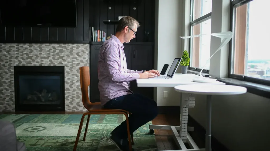 A man works at a laptop at a desk beside large windows, that can contribute to neck, shoulder and upper back tension.