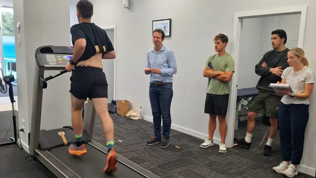 A runner undergoes treadmill gait analysis as physiotherapy staff observe their movement.